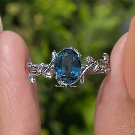 Blue oval-cut sapphire engagement ring with detailed leaf side mounts and silver prongs