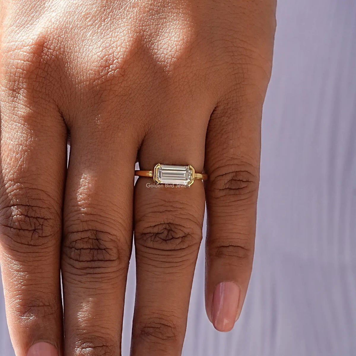 Emerald-cut east-west solitaire ring detail on hand, square prong setting