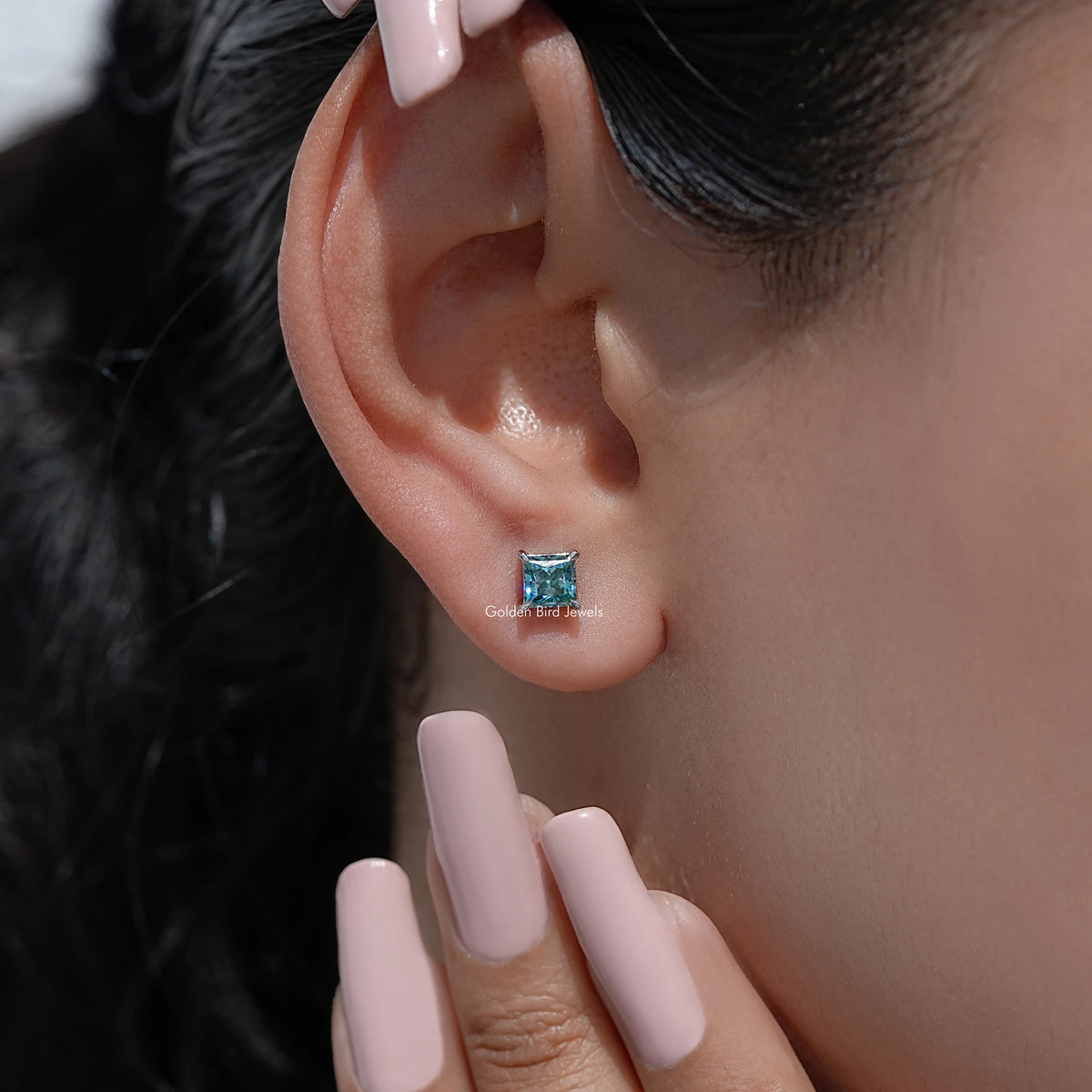 Blue princess-cut screw-back stud earrings on ear with manicured nails