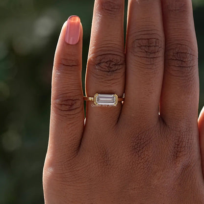 Emerald-cut east-west solitaire ring on finger, gold-tone prongs and slim band